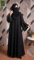 A woman wearing a black abaya with decorative gold chain trim on the sleeves and a matching black hijab, standing in front of a geometric tiled wall.