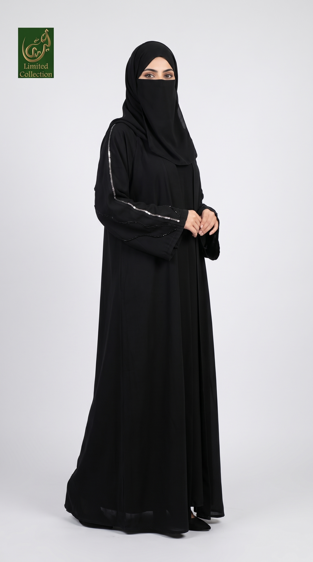 A woman modeling a black abaya and matching hijab against a light gray studio background. The abaya has subtle silver metallic trim along the sleeve edges, and a green 'Abayat Limited Collection' logo is visible in the top-left corner.