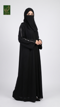 A woman modeling a black abaya and matching hijab against a light gray studio background. The abaya has subtle silver metallic trim along the sleeve edges, and a green 'Abayat Limited Collection' logo is visible in the top-left corner.