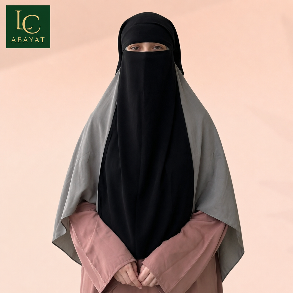 A woman wearing a black full-face niqab and a light gray shawl over a dusty pink long-sleeve top, standing against a soft peach background. The Abayat brand logo is visible in the top-left corner.