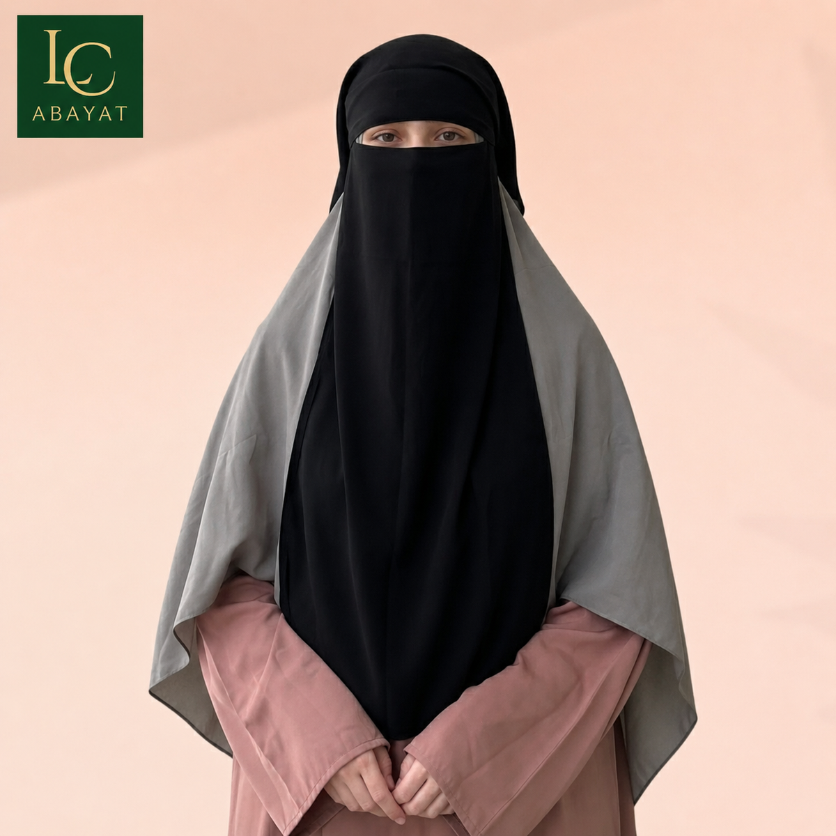 A woman wearing a black full-face niqab and a light gray shawl over a dusty pink long-sleeve top, standing against a soft peach background. The Abayat brand logo is visible in the top-left corner.
