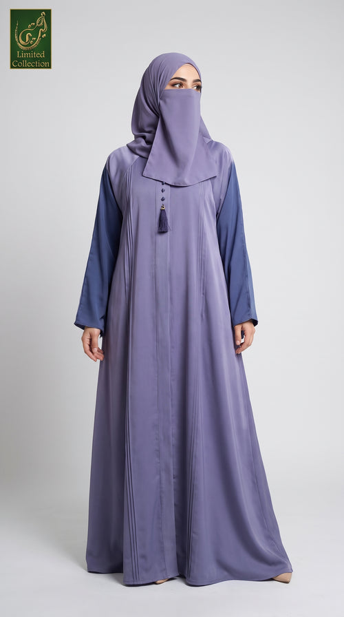 A woman modeling a modest long-sleeve dubai style pleated maxi abaya in a dusty lavender-purple color, paired with a matching hijab. The abaya features a contrasting darker blue-purple yoke at the shoulders and a small tassel detail at the neckline.