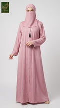 A woman wearing a dusty pink Dubai Style Pleated Maxi Abaya With Tassel Button in Premium Romantic Nida fabric with a matching face veil (niqab) and a small black tassel at the neckline, standing against a light gray studio background. A 'Abayat Limited Collection' logo is visible in the top-left corner.