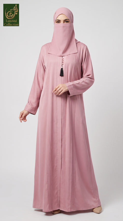 A woman wearing a dusty pink Dubai Style Pleated Maxi Abaya With Tassel Button in Premium Romantic Nida fabric with a matching face veil (niqab) and a small black tassel at the neckline, standing against a light gray studio background. A 'Abayat Limited Collection' logo is visible in the top-left corner.
