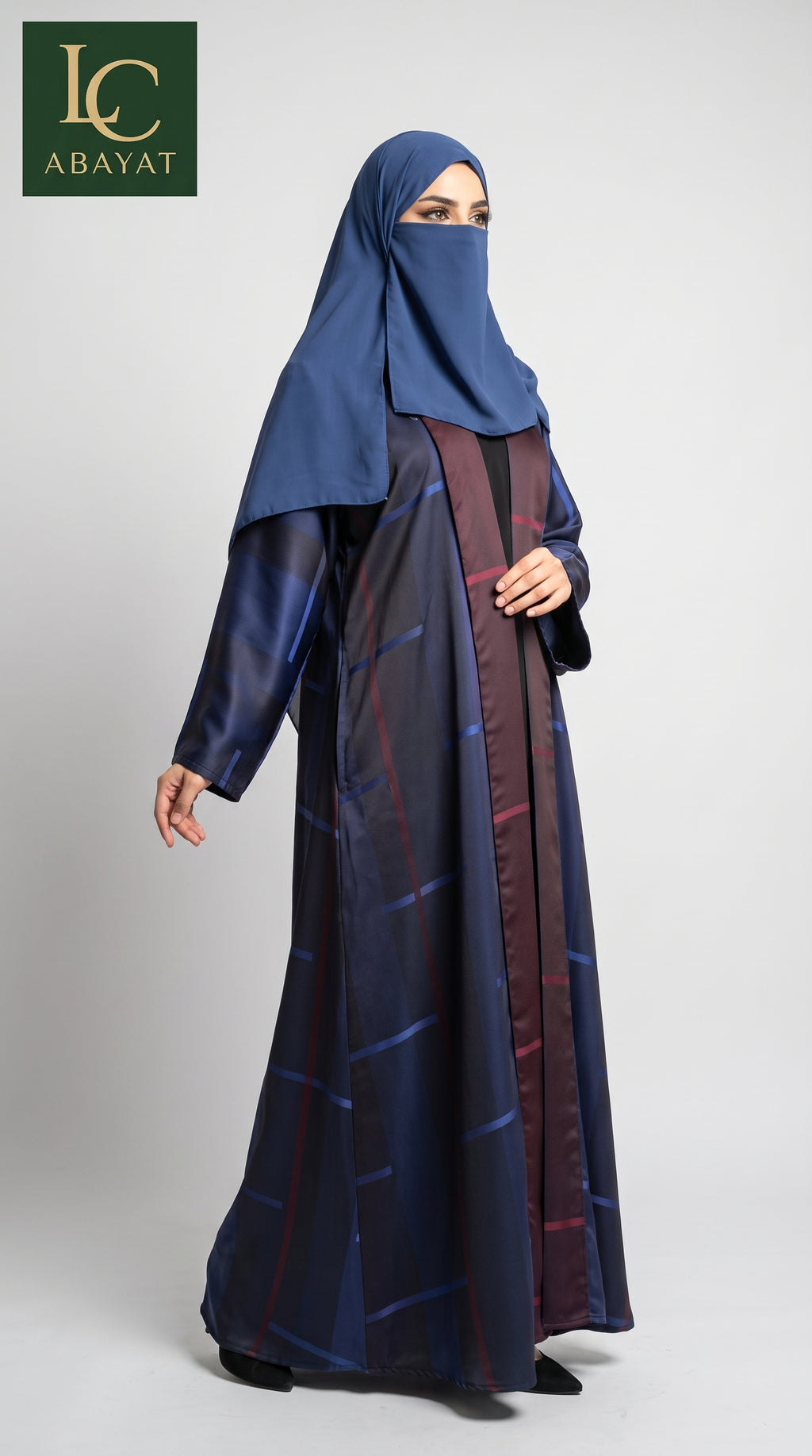 A woman wearing a modest blue abaya and matching hijab, standing against a light gray studio background. The abaya has a subtle grid pattern with darker blue and burgundy tones.