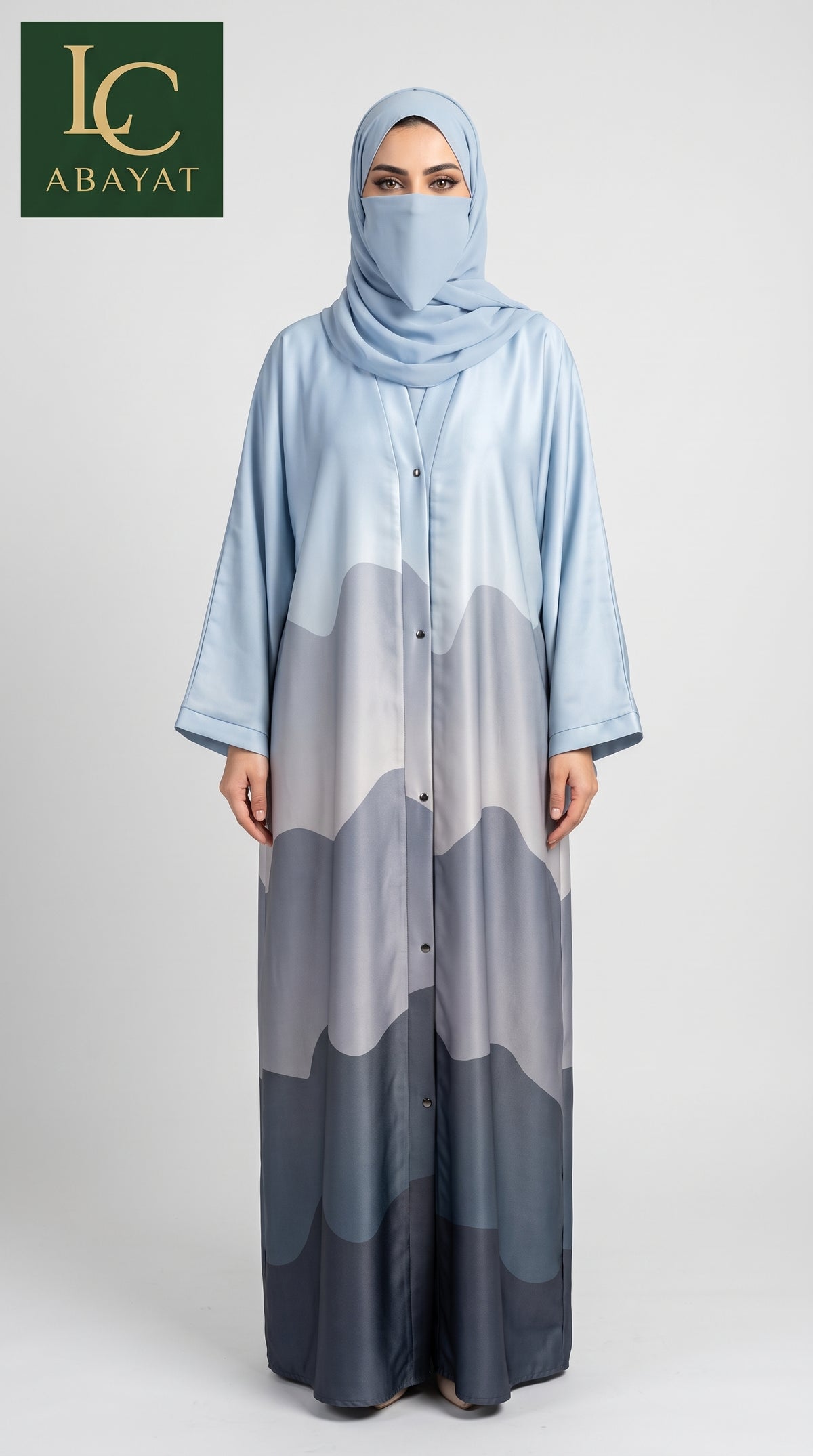 A woman modeling a light blue abaya with a mountain-patterned print in shades of gray and navy, paired with a matching light blue hijab. The logo for 'LC ABAYAT' is visible in the top left corner.