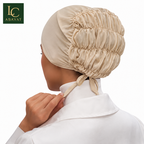 A beige satin-like head covering or elastic hijab cap with gathered ruching and a tie at the back, shown on a model from the back. The model wears a white collared shirt.