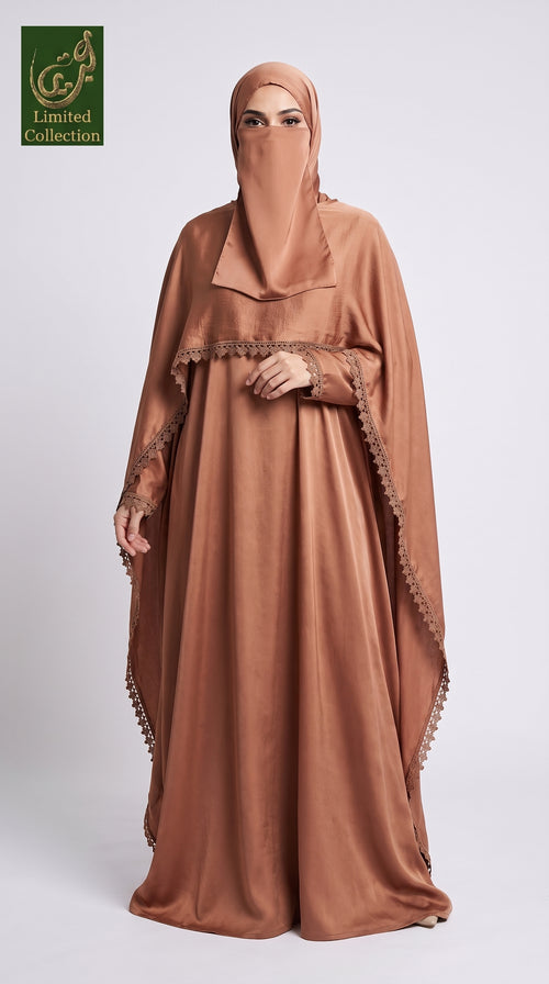 A woman modeling a modest brown maxi abaya with a overlay shrug with a matching hijab and face veil, standing against a light gray studio background. The abaya has draped sleeves and decorative trim along the edges.