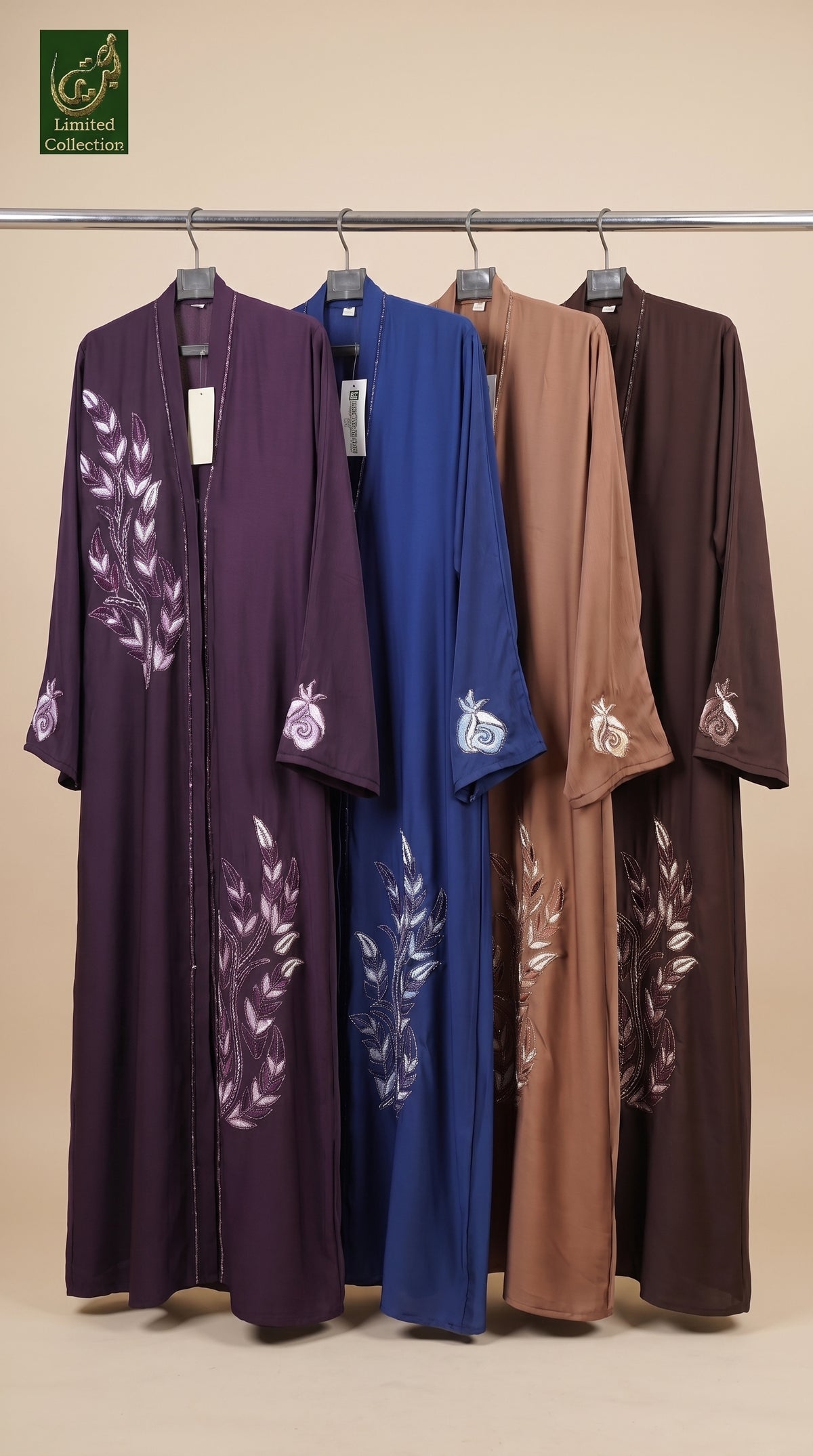 Four long open-front coloful stonework abayas hanging on a metal rack against a beige background, shown in purple, blue, tan, and brown, each with white embroidered leaf motifs on the front and sleeves.