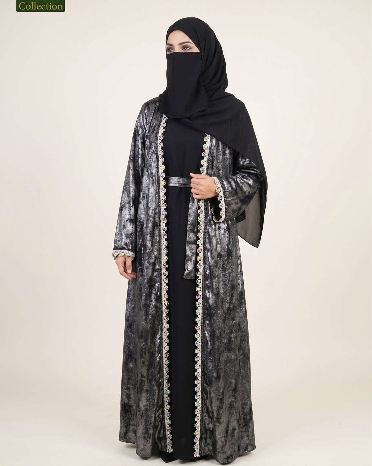 A woman modeling a long black abaya with a shimmering silver-gray outer layer, black embroidered trim, a matching black headscarf, and a black face veil against a plain beige studio background.