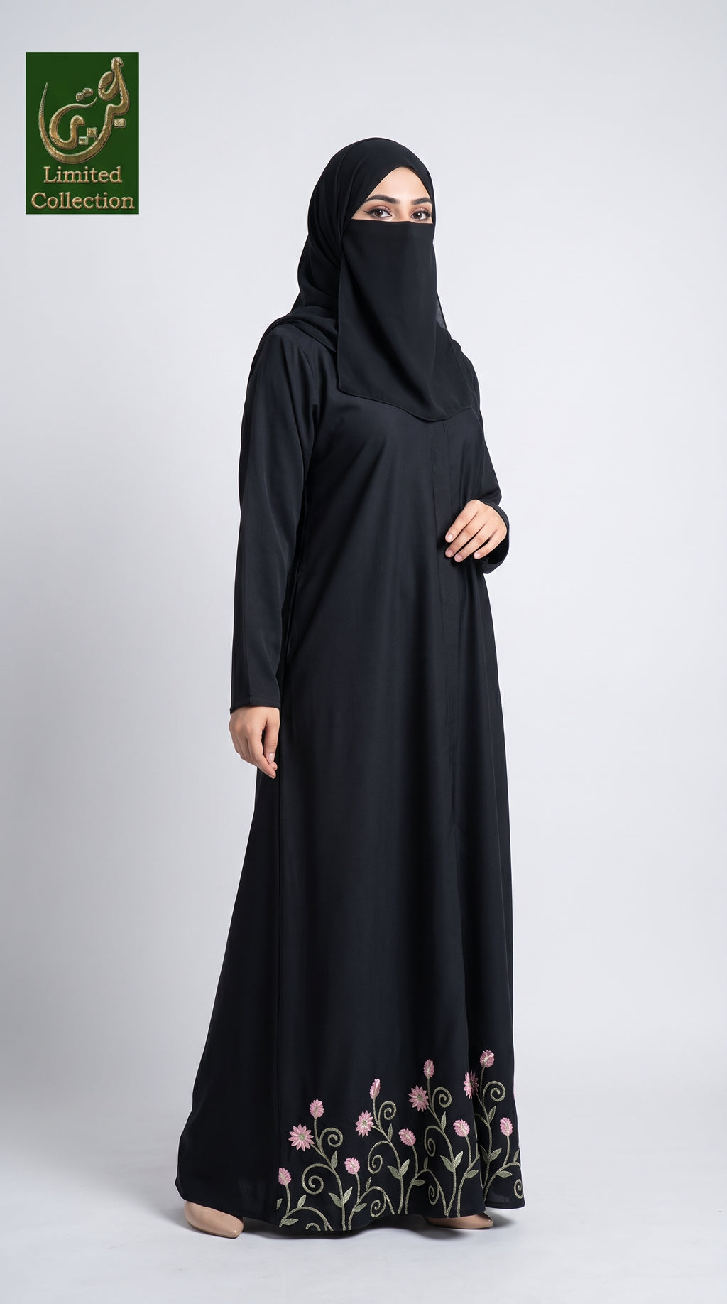 A woman wearing a black long-sleeve abaya with a matching black hijab and a face veil, standing against a light gray studio background. The abaya features pink floral embroidery and green vine detailing near the hem.