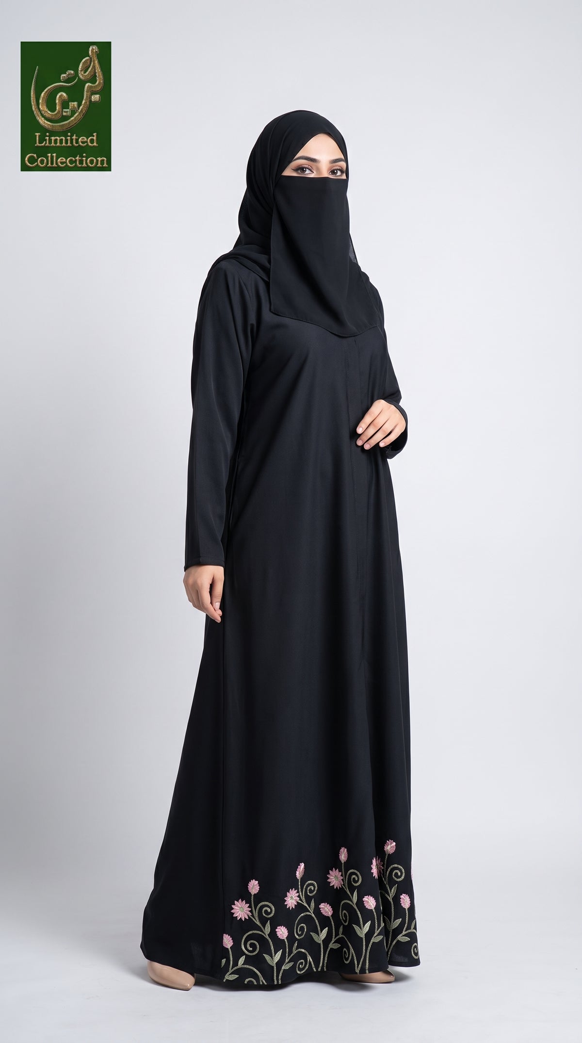 A woman wearing a black long-sleeve abaya with a matching black hijab and a face veil, standing against a light gray studio background. The abaya features pink floral embroidery and green vine detailing near the hem.