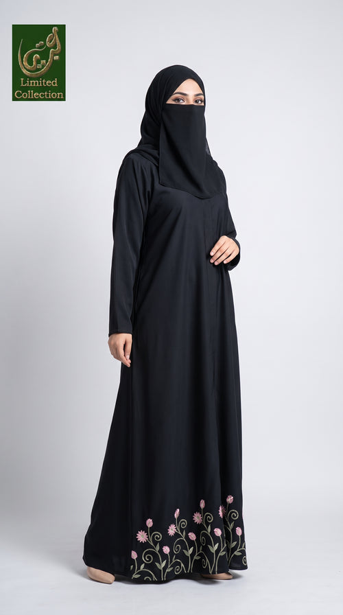 A woman wearing a black long-sleeve abaya with a matching black hijab and a face veil, standing against a light gray studio background. The abaya features pink floral embroidery and green vine detailing near the hem.