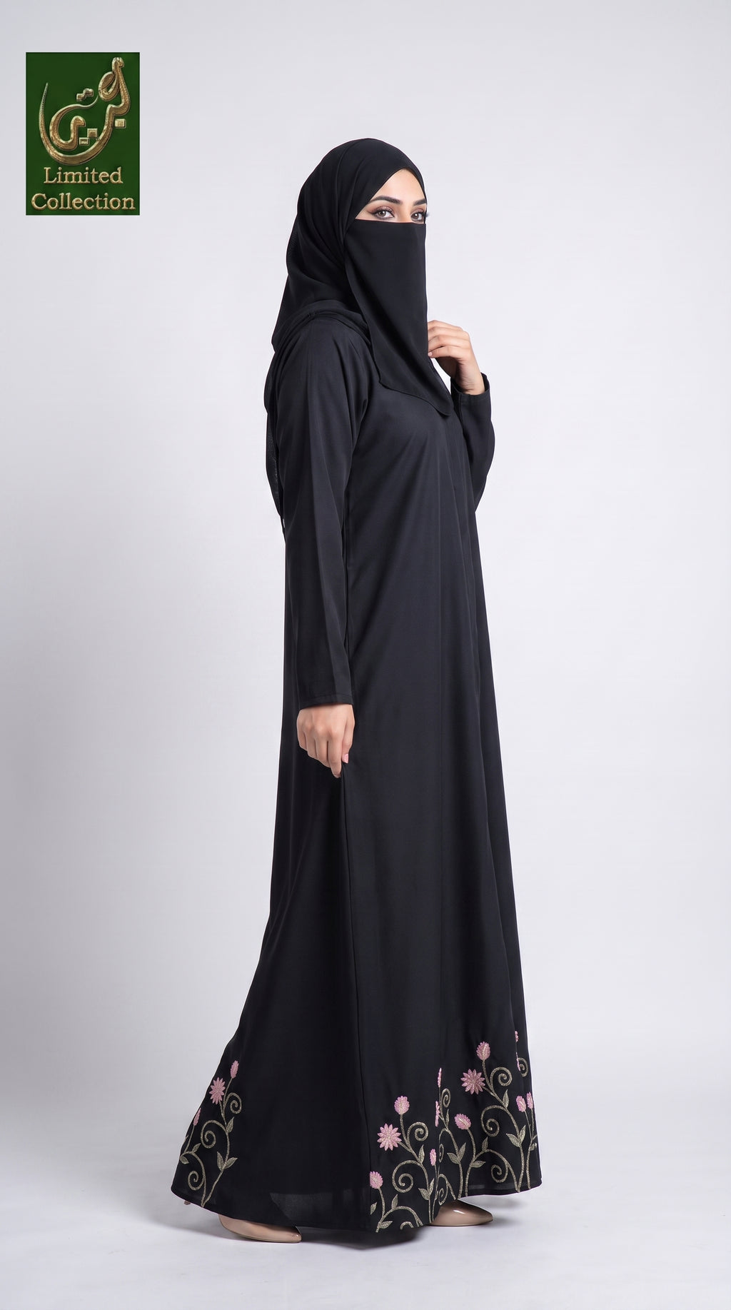 A woman modeling a black abaya and matching hijab with pink and green floral embroidery near the hem, photographed against a light gray studio background. A green 'Limited Collection' logo appears in the top-left corner.