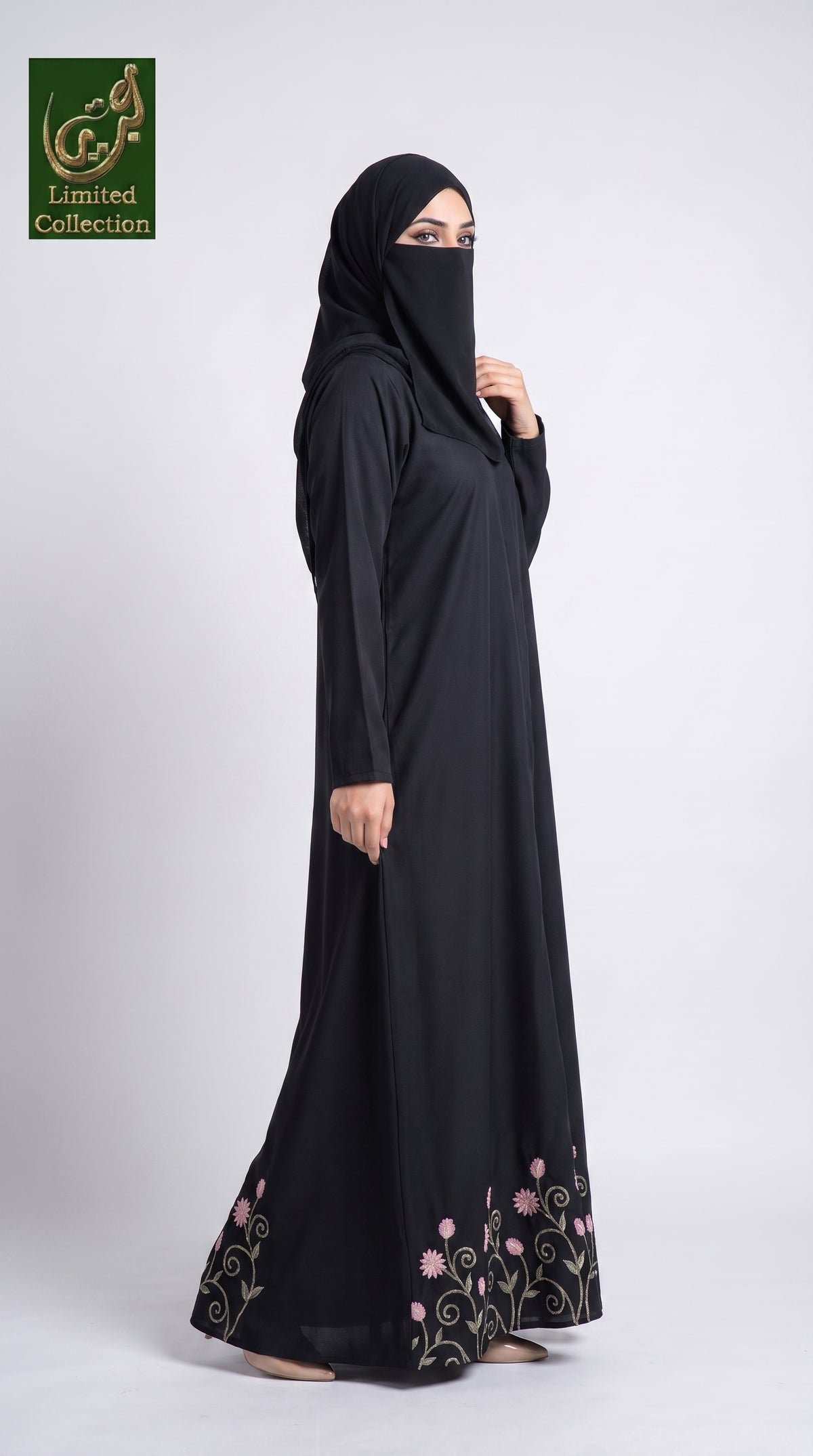 A woman modeling a black abaya and matching hijab with pink and green floral embroidery near the hem, photographed against a light gray studio background. A green 'Limited Collection' logo appears in the top-left corner.