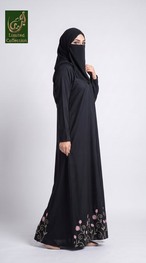 A woman modeling a black abaya and matching hijab with pink and green floral embroidery near the hem, photographed against a light gray studio background. A green 'Limited Collection' logo appears in the top-left corner.