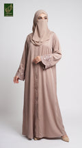 A woman modeling a long taupe-beige abaya with floral lace sleeve embroidery and a matching hijab, standing against a light gray studio background. A green 'Lumiere Collection' logo is visible in the top-left corner.