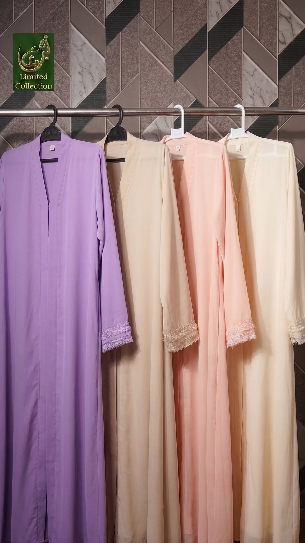 Four long modest plain abaya hanging on a rack against a geometric tiled wall. The leftmost gown is lavender purple, and the others are cream, light beige, blush pink, and off-white.