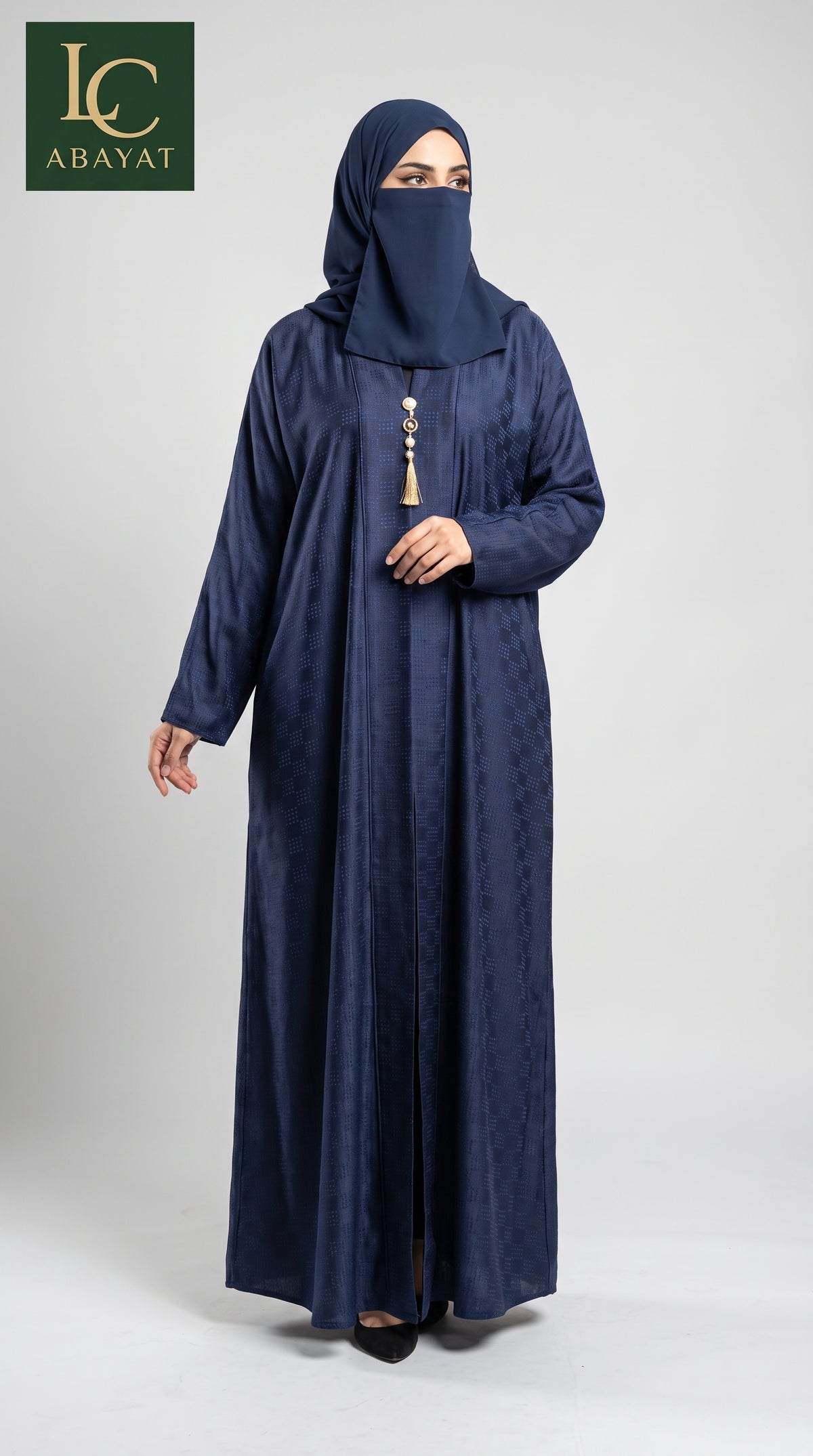 A woman wearing a navy blue abaya and matching hijab with a decorative tassel at the neckline, standing against a light gray studio background. The LC Abayat brand logo is visible in the top left corner.