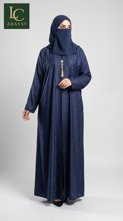A woman wearing a navy blue abaya and matching hijab with a decorative tassel at the neckline, standing against a light gray studio background. The LC Abayat brand logo is visible in the top left corner.