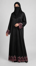 A woman wearing a long black abaya with pink and red floral embroidery on the cuffs and hem, paired with a matching black hijab, photographed against a light gray studio background.