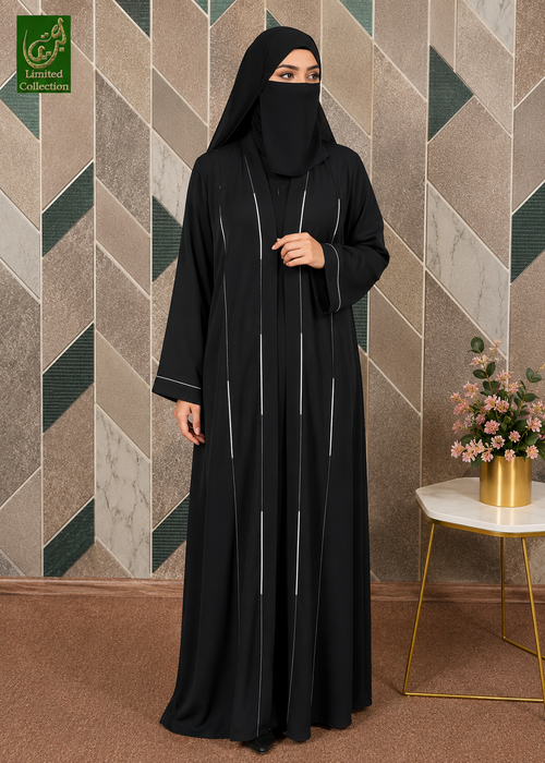A woman wearing a black Korean Nida piping work abaya with thin vertical silver-white stripes and a matching black hijab, standing in front of a geometric patterned wall. A small side table with pink flowers is on the right, and a green 'Luxeen Collection' logo is in the top-left corner.