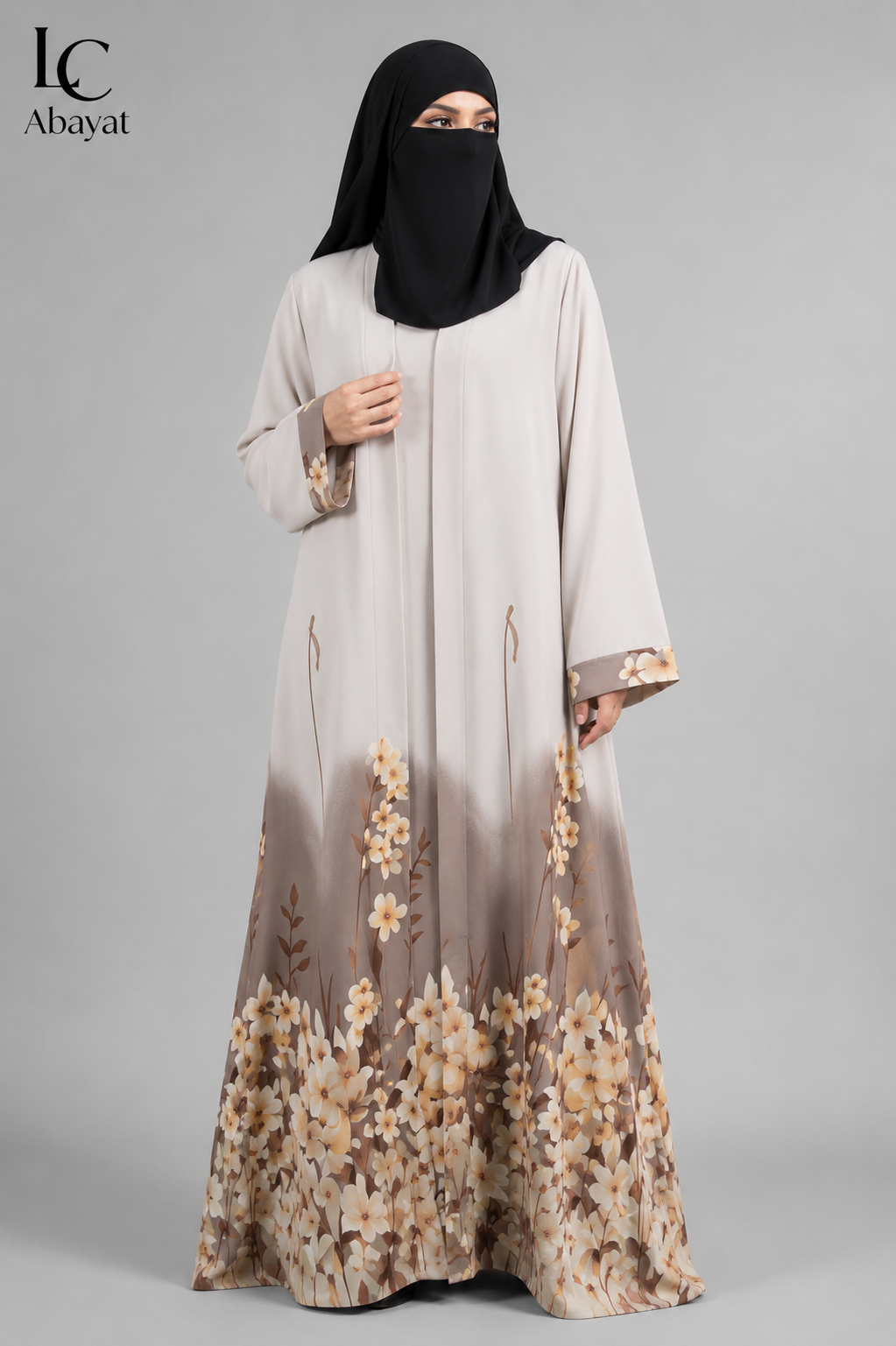 A woman wearing a long front open floral border printed abaya with a black hijab, standing against a plain gray studio background. The abaya is light beige at the top and fades into taupe and brown at the bottom, with a floral print concentrated near the hem.