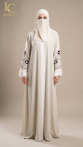 A woman wearing a long cream abaya with purple embroidered details on the sleeves and a matching cream hijab, standing against a solid beige studio background. The LC Abayat brand logo is visible in the top left corner.
