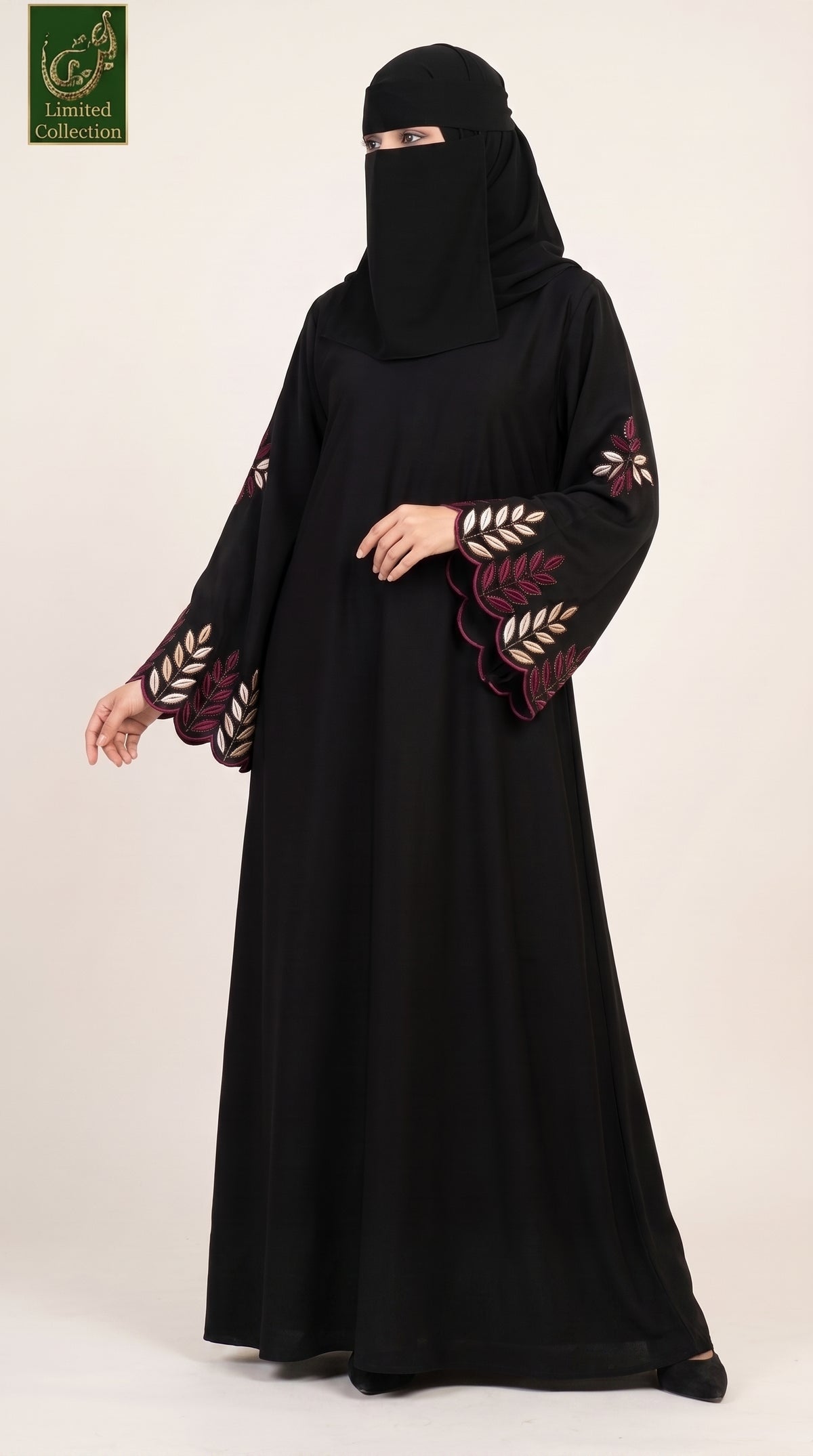 A woman wearing a black abaya with embroidered leaf patterns on the sleeves and a matching black face covering, standing against a plain light beige studio background. A green 'Limited Collection' logo is visible in the top-left corner.