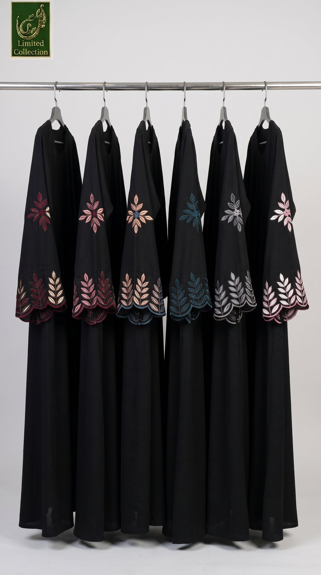 Six black abayas hanging on a metal rack against a light gray background, each decorated with colorful embroidered floral and leaf patterns in pink, blue, and silver. A green 'Limited Collection' logo is visible in the top left corner.