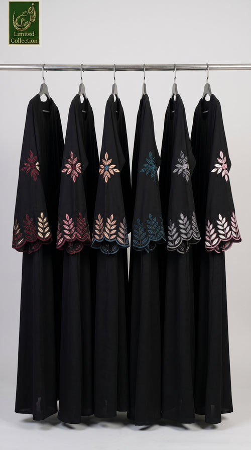 Six black abayas hanging on a metal rack against a light gray background, each decorated with colorful embroidered floral and leaf patterns in pink, blue, and silver. A green 'Limited Collection' logo is visible in the top left corner.