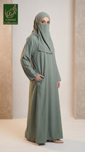 A woman modeling a sage green abaya with a matching hijab, standing in a studio setting with textured beige walls. The abaya has long sleeves and side pockets, and a logo for 'Arab Limited Collection' is visible in the top left corner.