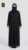A woman wearing a long black abaya and matching black hijab stands against a light gray studio background. A small green logo appears in the top-left corner.