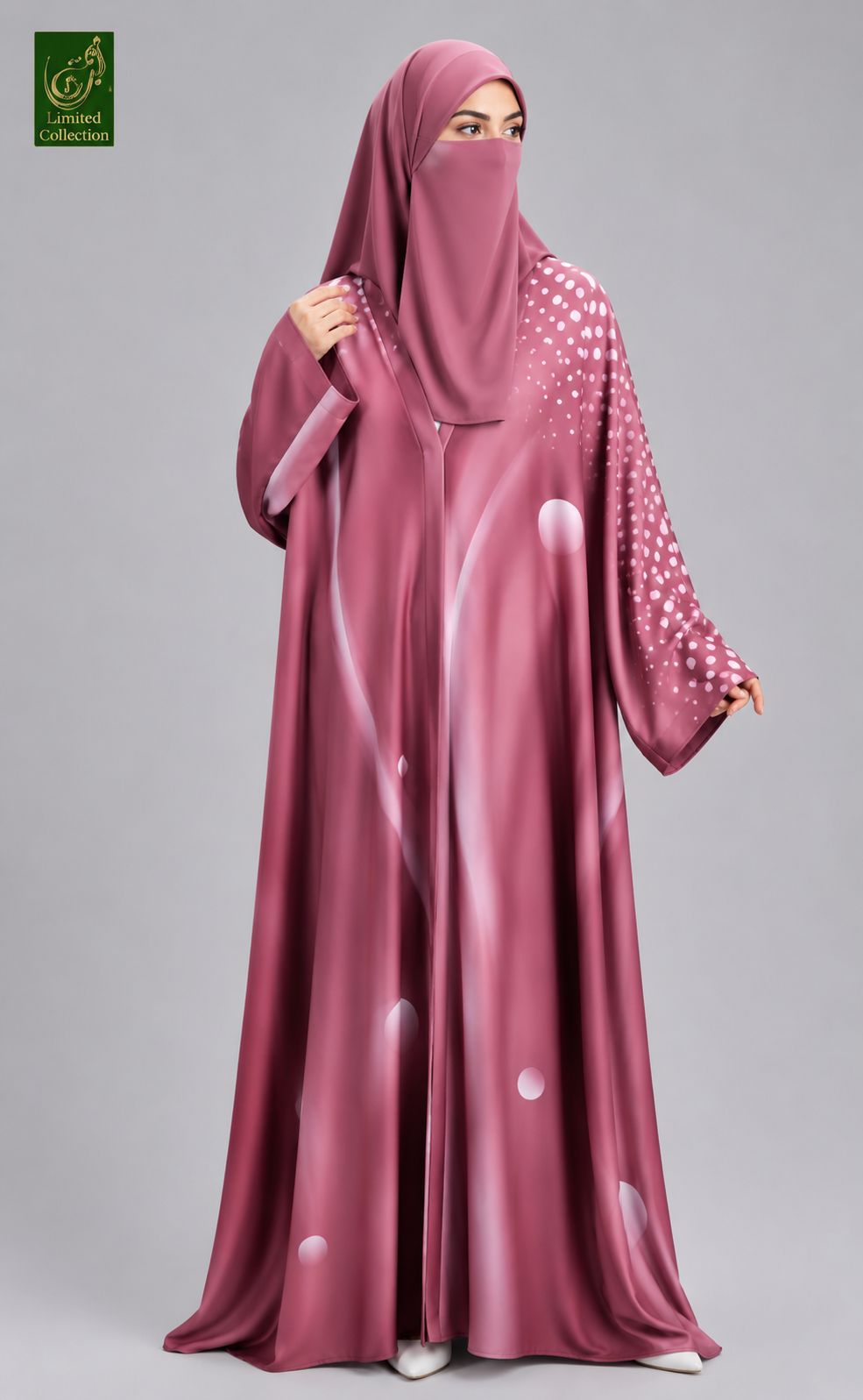 A woman wearing a long, flowing magenta-pink abaya and matching hijab with white circular dot patterns on the sleeves, photographed against a plain light gray studio background.