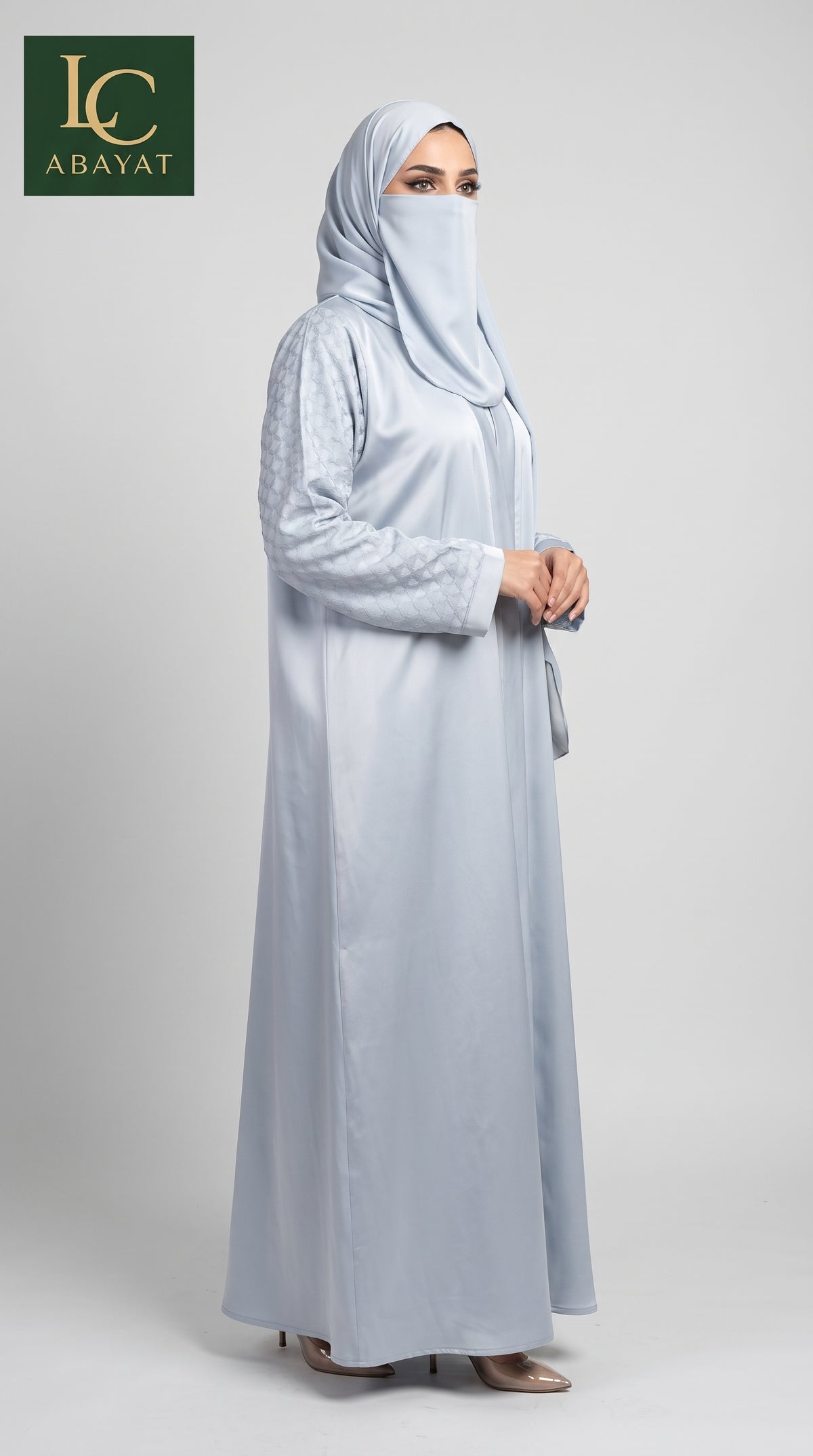A woman modeling a light blue-gray abaya and matching hijab against a neutral studio background, with the LC Abayat logo in the top-left corner.
