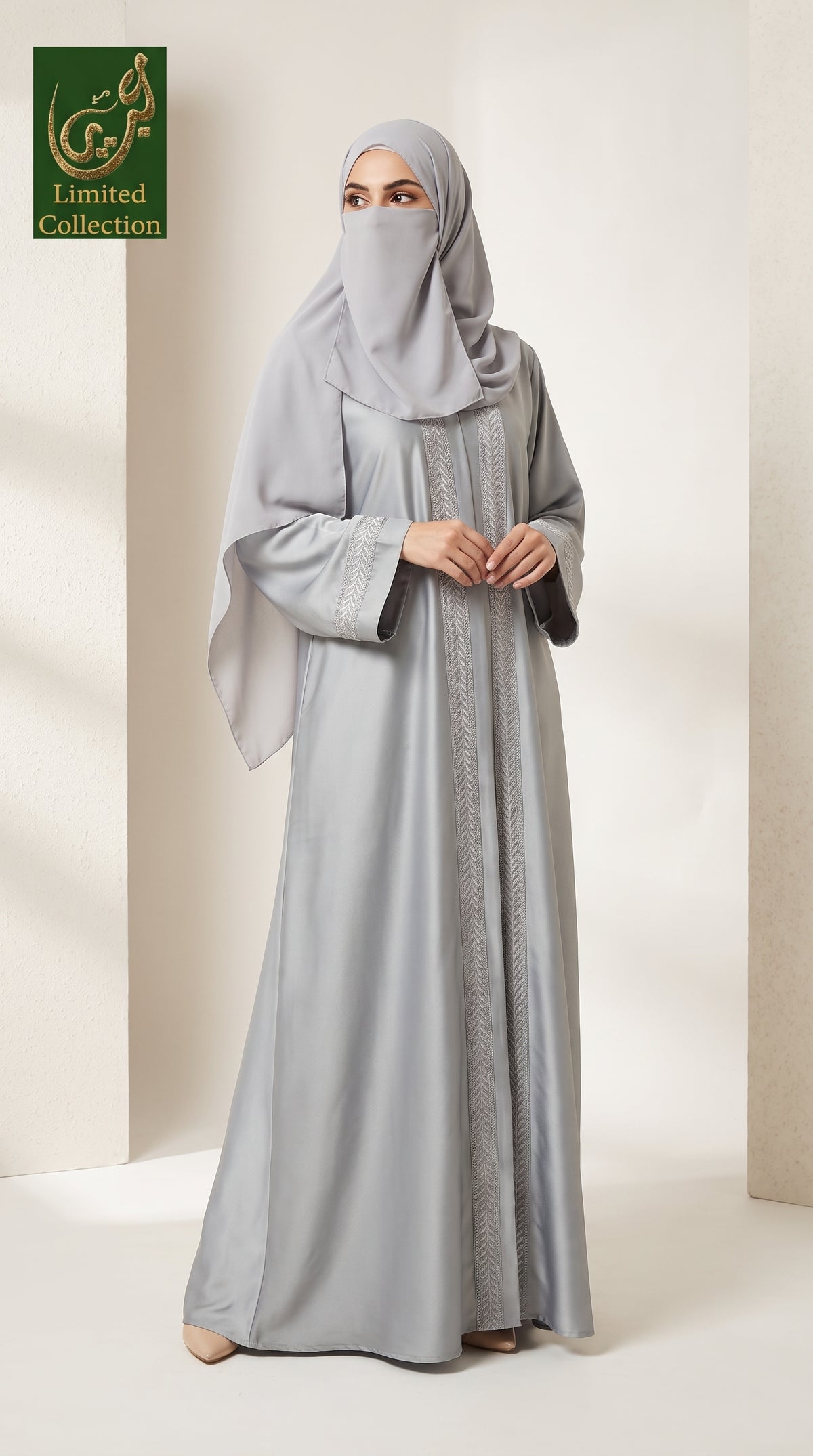 A woman wearing a light gray abaya and matching hijab stands in a studio setting. The abaya has long sleeves, a modest silhouette, and subtle vertical embroidery down the front center.