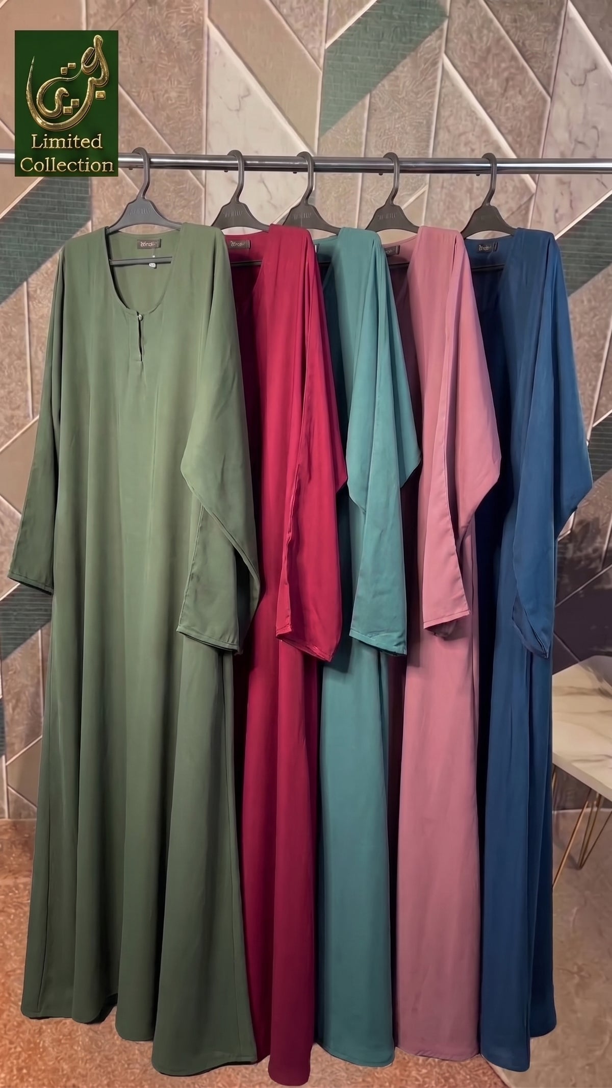 Five long modest plain maxi front-closed abayas hanging on a metal rack against a patterned wall. The garments are shown in olive green, burgundy, teal, dusty pink, and navy blue.