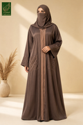 A woman wearing a long brown front open abaya with pintex pocksts and a matching hijab, standing in a studio setting with a beige background and a decorative vase of dried grass on the left. The abaya features a central front opening with a contrasting brown panel and quilted patch pockets.