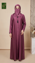 A woman wearing a long, floor-length mauve-purple abaya with a matching hijab and face veil, standing in a studio with a beige background and a decorative plant on the right.