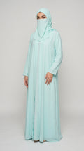 A woman modeling a long, flowing abaya in a light mint green color, paired with a matching hijab and face veil, against a plain light gray studio background.