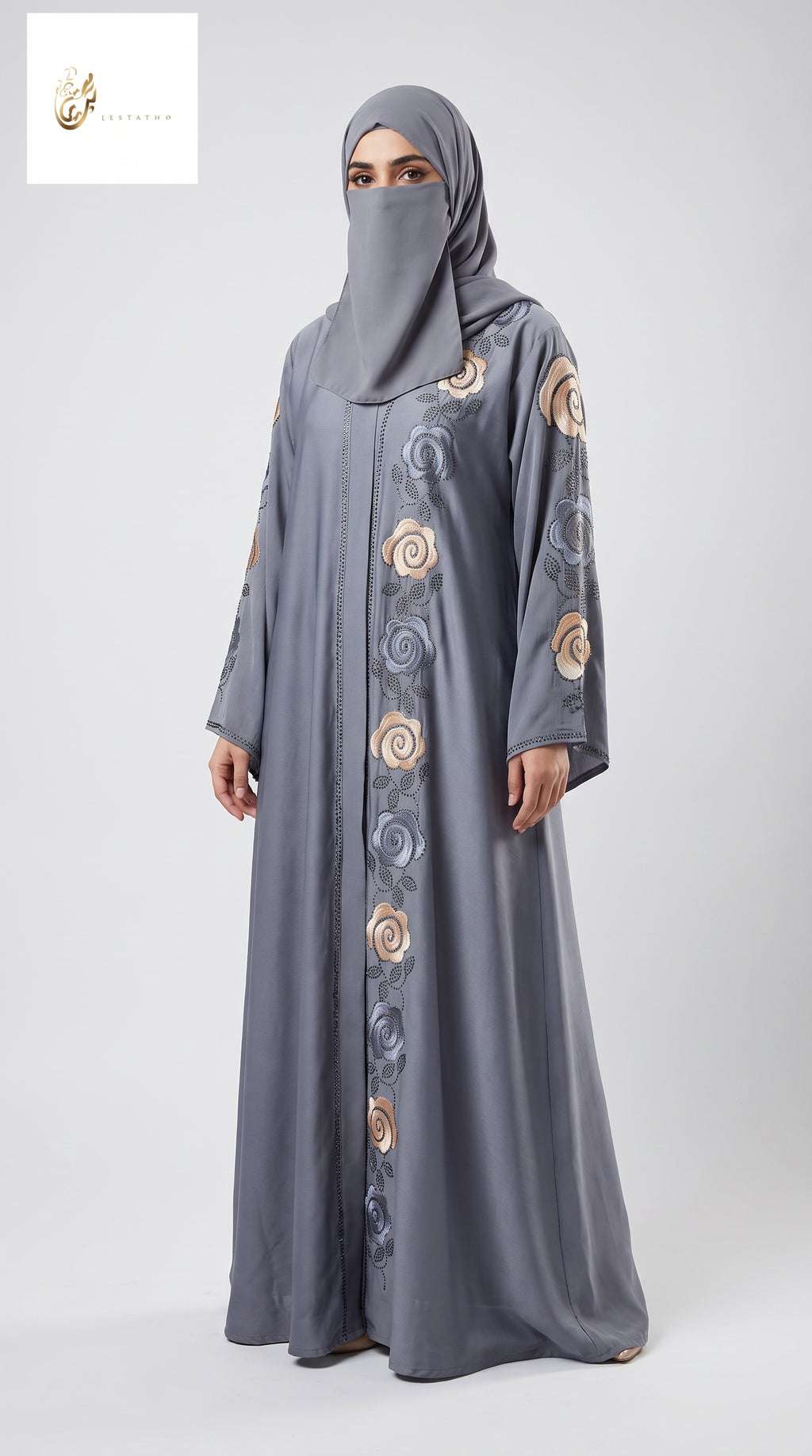 A woman wearing a long gray abaya with matching hijab, featuring circular embroidered patterns in beige, gold, and blue-gray down the front and on the sleeves, photographed against a light gray studio background.