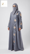 A woman wearing a long gray abaya with matching hijab, featuring circular embroidered patterns in beige, gold, and blue-gray down the front and on the sleeves, photographed against a light gray studio background.