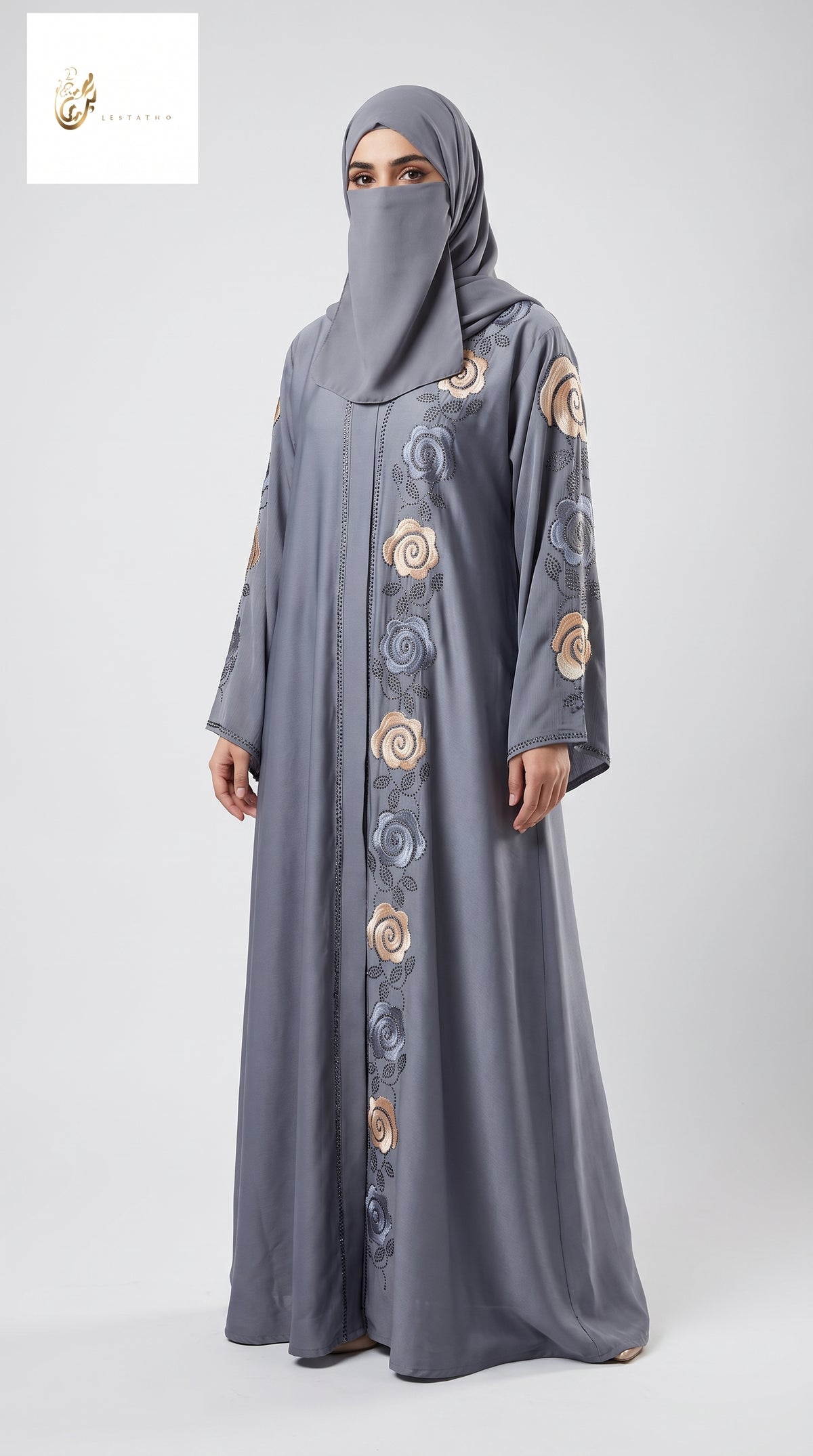 A woman wearing a long gray abaya with matching hijab, featuring circular embroidered patterns in beige, gold, and blue-gray down the front and on the sleeves, photographed against a light gray studio background.