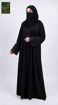 A woman wearing a full-length black abaya with stonework lace embroidered sleeves and a matching black hijab, standing against a light gray studio background. A green 'Abayat Limited Collection' logo appears in the top-left corner.