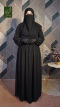 A woman wearing a full-length gathered maxi black abaya with long sleeves, a matching black face veil, and black gloves, standing in front of a geometric patterned wall. A small side table with flowers is visible on the right, and a green 'Limited Collection' logo appears in the top-left corner.