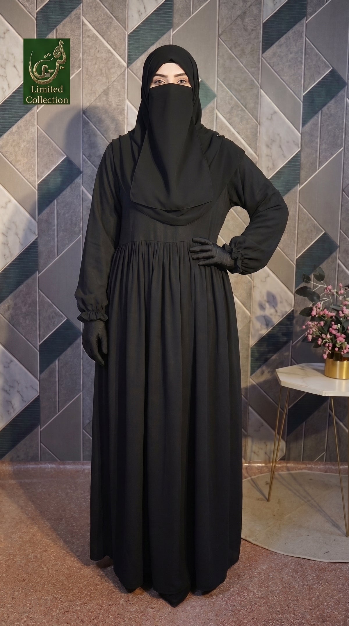 A woman wearing a long black maxi abaya dress with long sleeves, a matching black head covering, and black gloves, standing in front of a geometric patterned wall. A small side table with flowers is visible on the right, and a logo watermark appears in the top left corner.