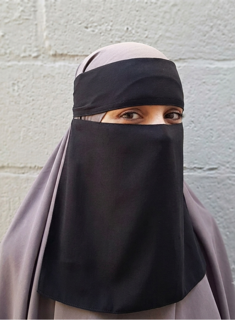 A woman wearing a black niqab/face veil and a light gray hijab stands against a light gray concrete block wall.
