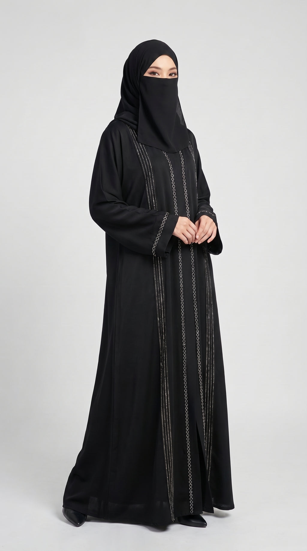 A woman wearing a long black abaya with vertical silver metallic embroidery and a matching black hijab, standing against a light gray studio background.