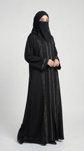 A woman wearing a long black abaya with vertical silver metallic embroidery and a matching black hijab, standing against a light gray studio background.