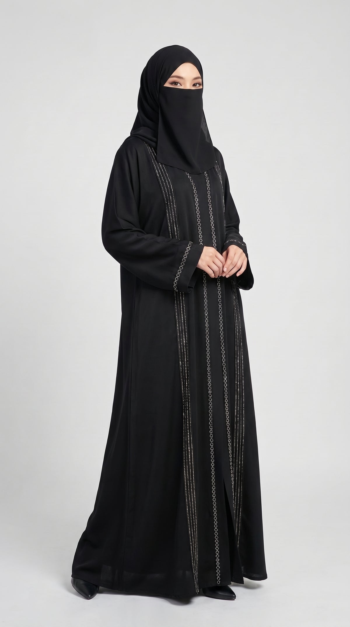 A woman wearing a long black abaya with vertical silver metallic embroidery and a matching black hijab, standing against a light gray studio background.